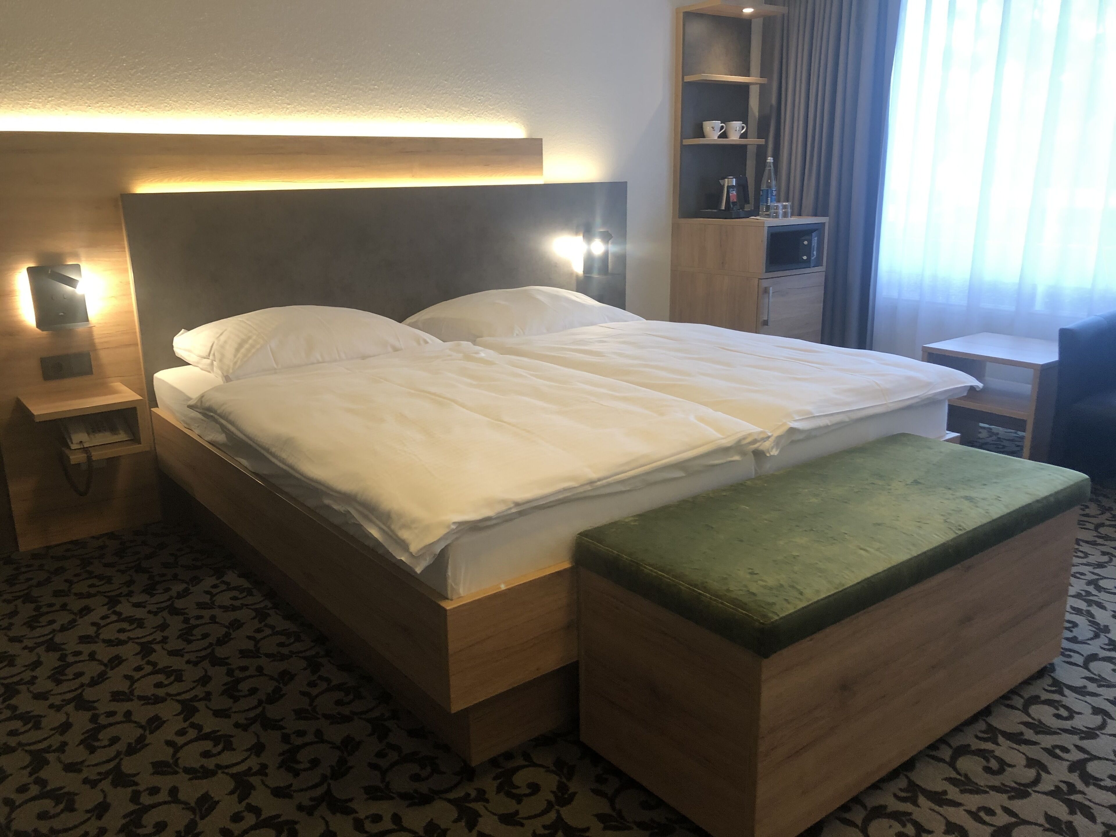 Habitación de hotel con una cama grande, una mesita de noche con lámpara, un banco de madera, un estante con tazas y una botella, y una alfombra estampada.