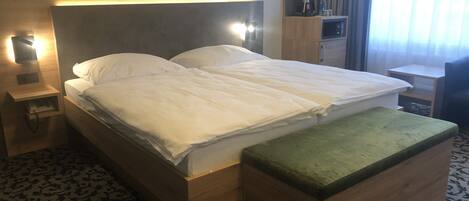 Habitación de hotel con una cama grande, una mesita de noche con lámpara, un banco de madera, un estante con tazas y una botella, y una alfombra estampada.