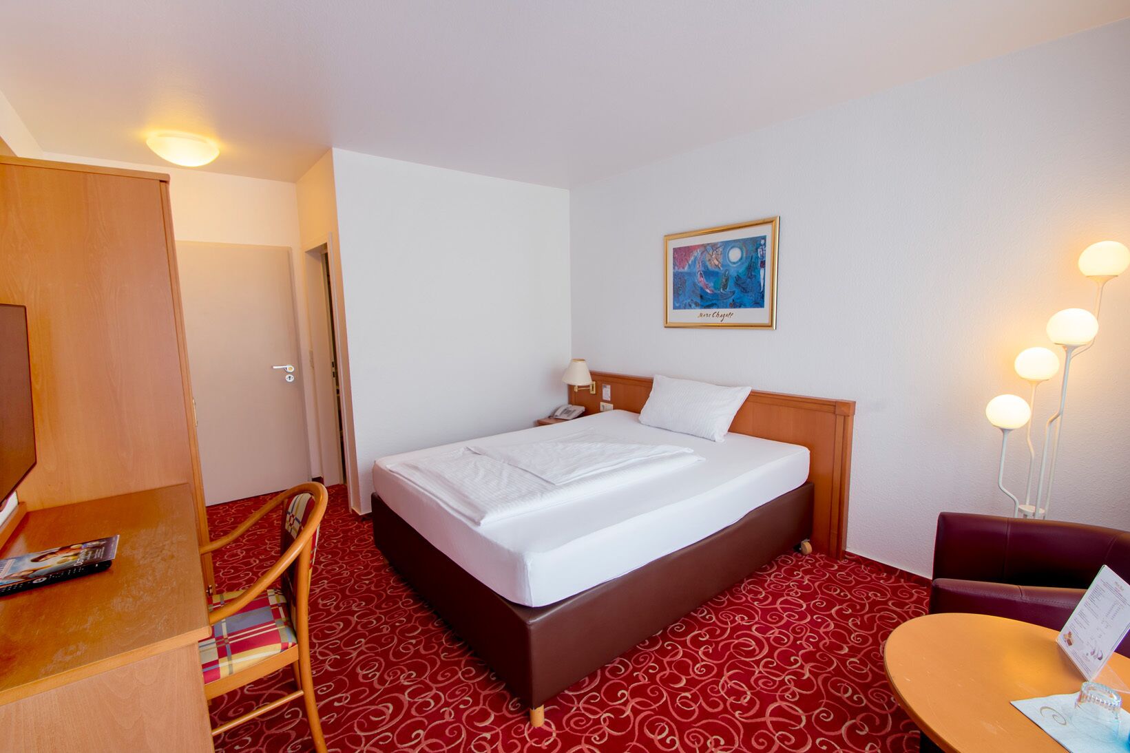 Una habitación de hotel con una cama, un escritorio con una silla, una mesita y una lámpara de pared.