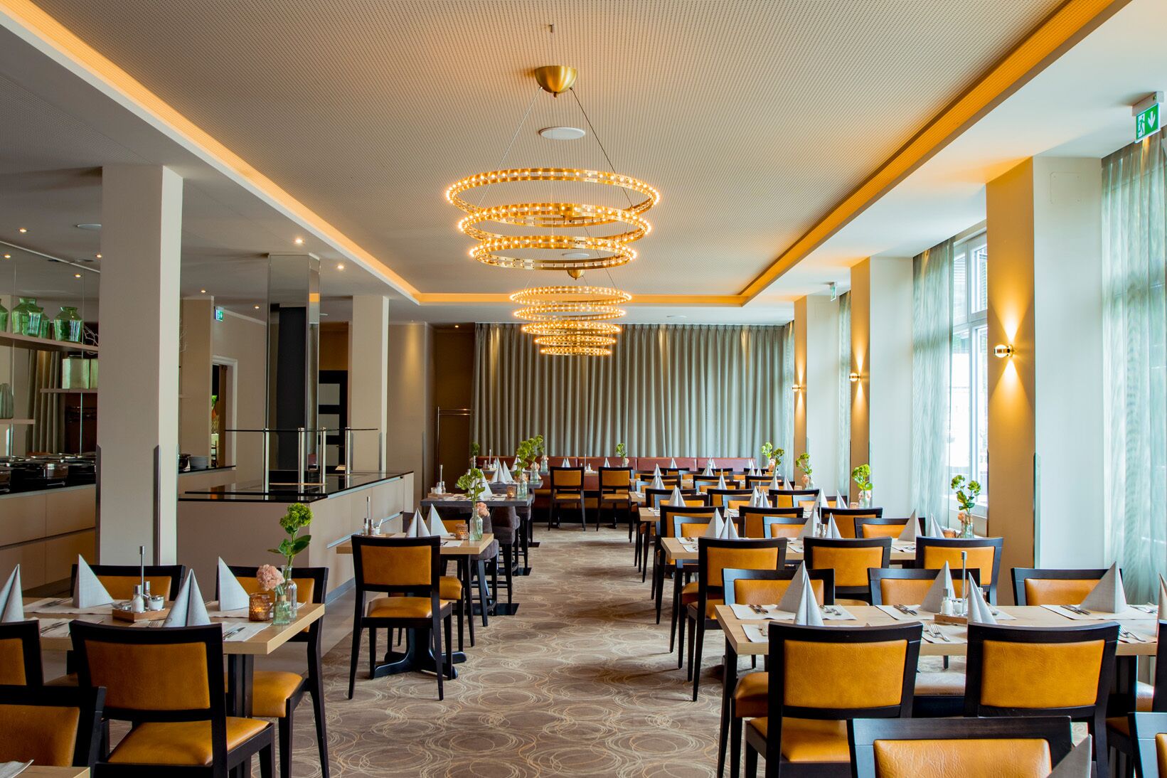 A spacious hotel dining area with multiple tables and chairs, a central chandelier, and large windows.