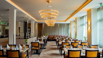 A spacious hotel dining area with multiple tables and chairs, a central chandelier, and large windows.