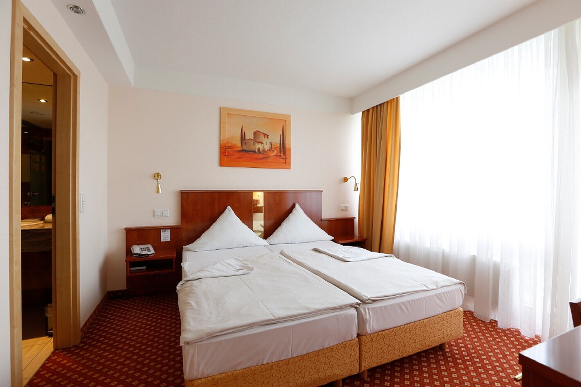 Quarto de hotel com uma cama de casal, mesa de cabeceira com luminária, quadro na parede e janela com cortinas.