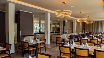 A spacious restaurant with multiple dining tables and chairs, a central bar area, and modern lighting fixtures.