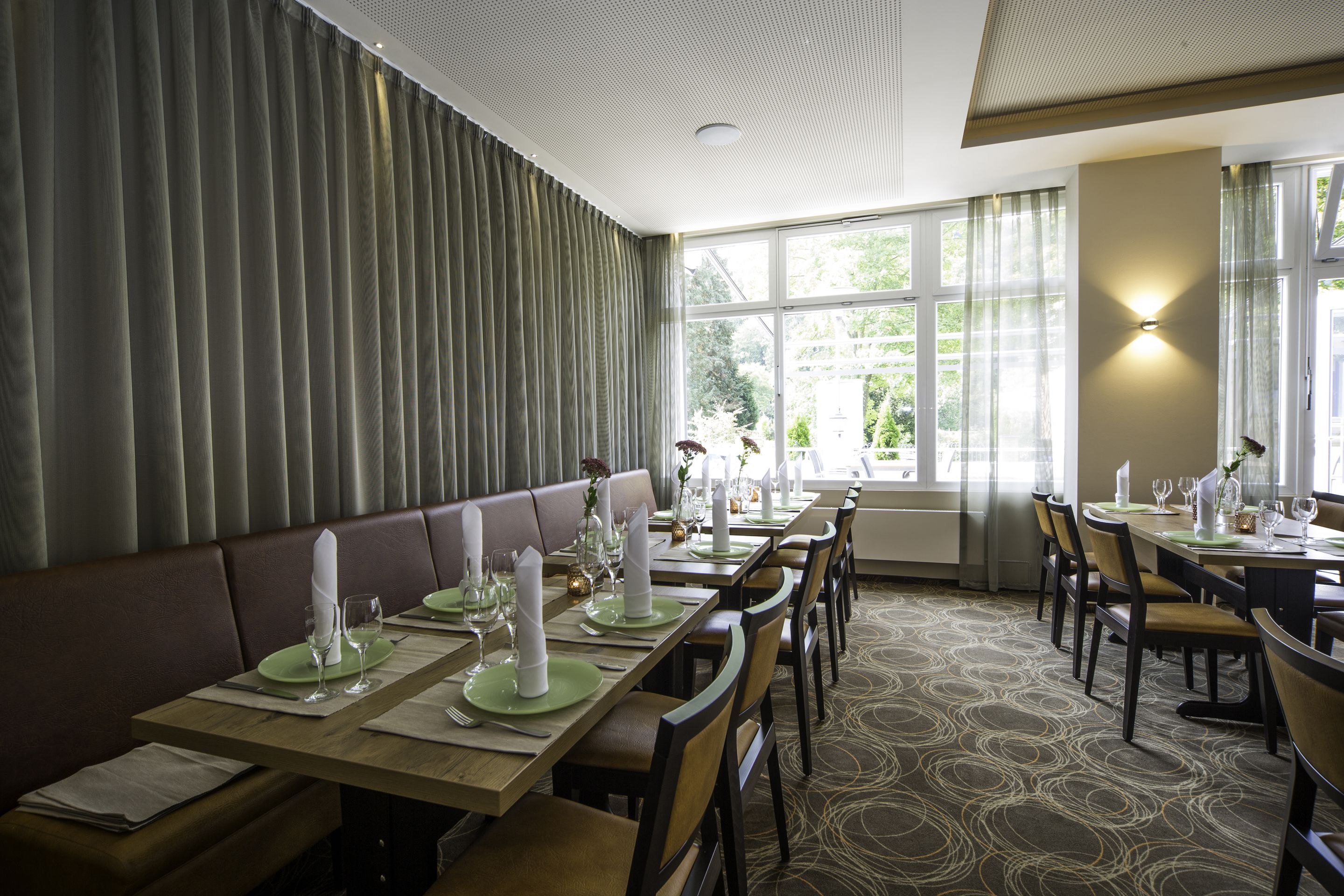 Un restaurante con una larga mesa de comedor dispuesta para una comida, asientos acolchados y ventanas amplias con cortinas.