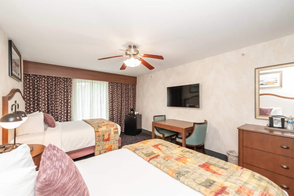A hotel room with two beds, a desk, a chair, a television, and a ceiling fan.