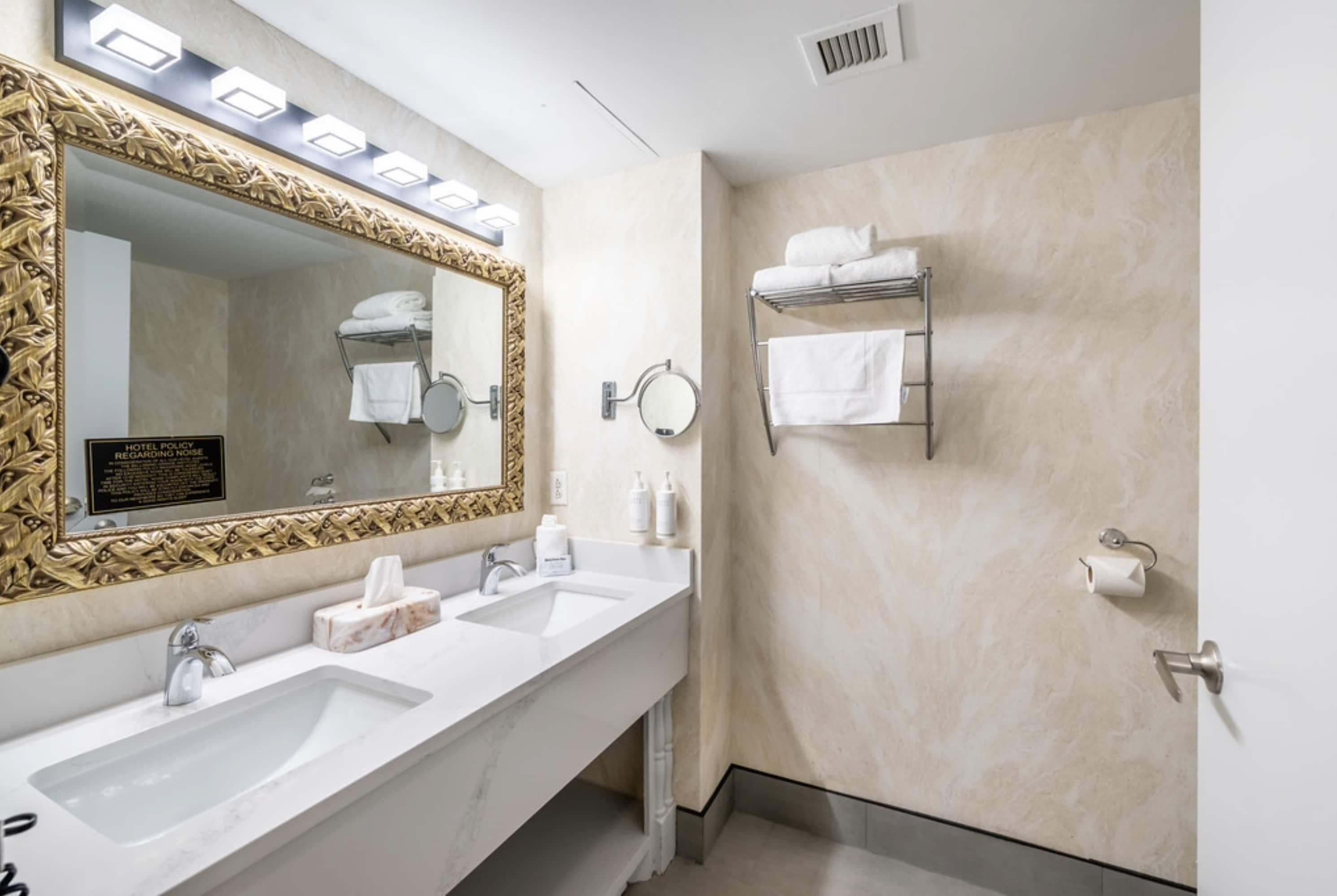 Une salle de bain d’hôtel avec deux lavabos, un grand miroir avec un cadre décoratif, des porte-serviettes et un panneau indiquant les règles concernant le niveau sonore.