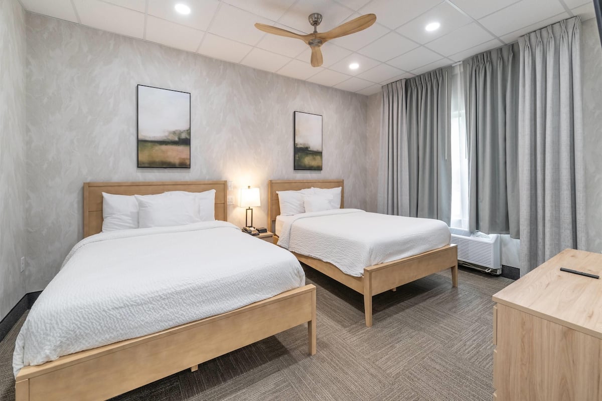 A hotel room with two beds, a ceiling fan, artwork on the walls, and a window with curtains.