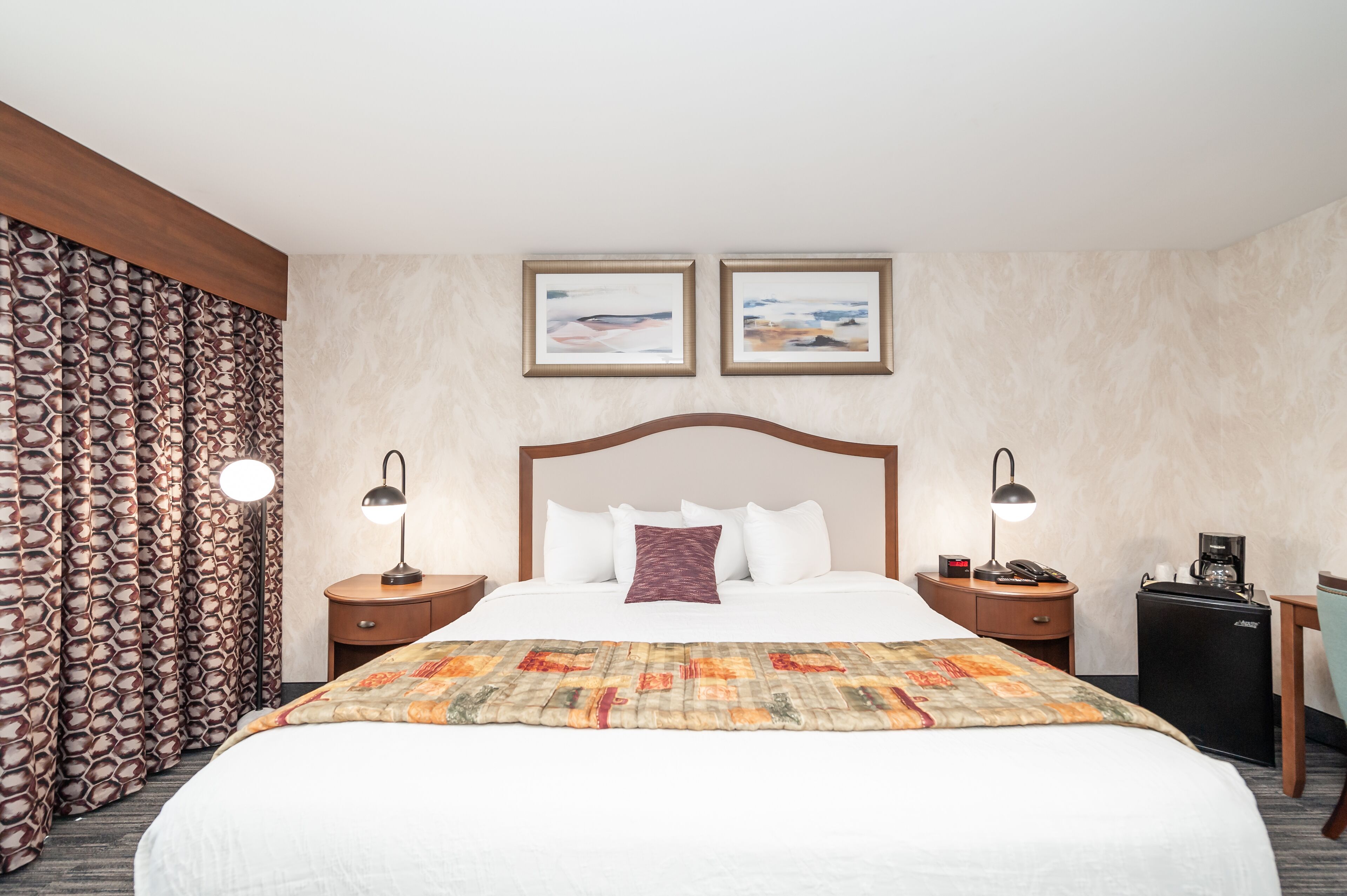 Ein Hotelzimmer mit einem großen Bett, zwei Nachttischen mit Lampen, einem kleinen Schreibtisch mit Stuhl, einer Kaffeemaschine und zwei gerahmten Bildern an der Wand.