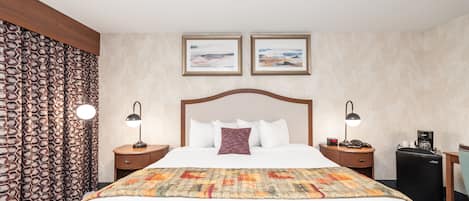Een hotelkamer met een groot bed, twee nachtkastjes met lampen, een klein bureau met stoel, een koffiezetapparaat en twee ingekaderde foto’s aan de muur.