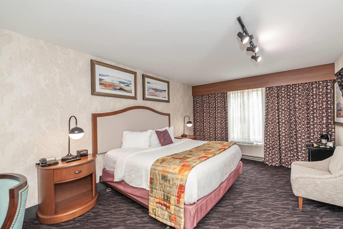A hotel room with a large bed, a nightstand with a lamp, a chair, and a desk with a coffee machine.