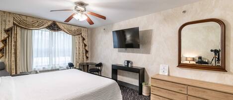 A hotel room with a bed, a desk, a TV, a mirror, and a ceiling fan.