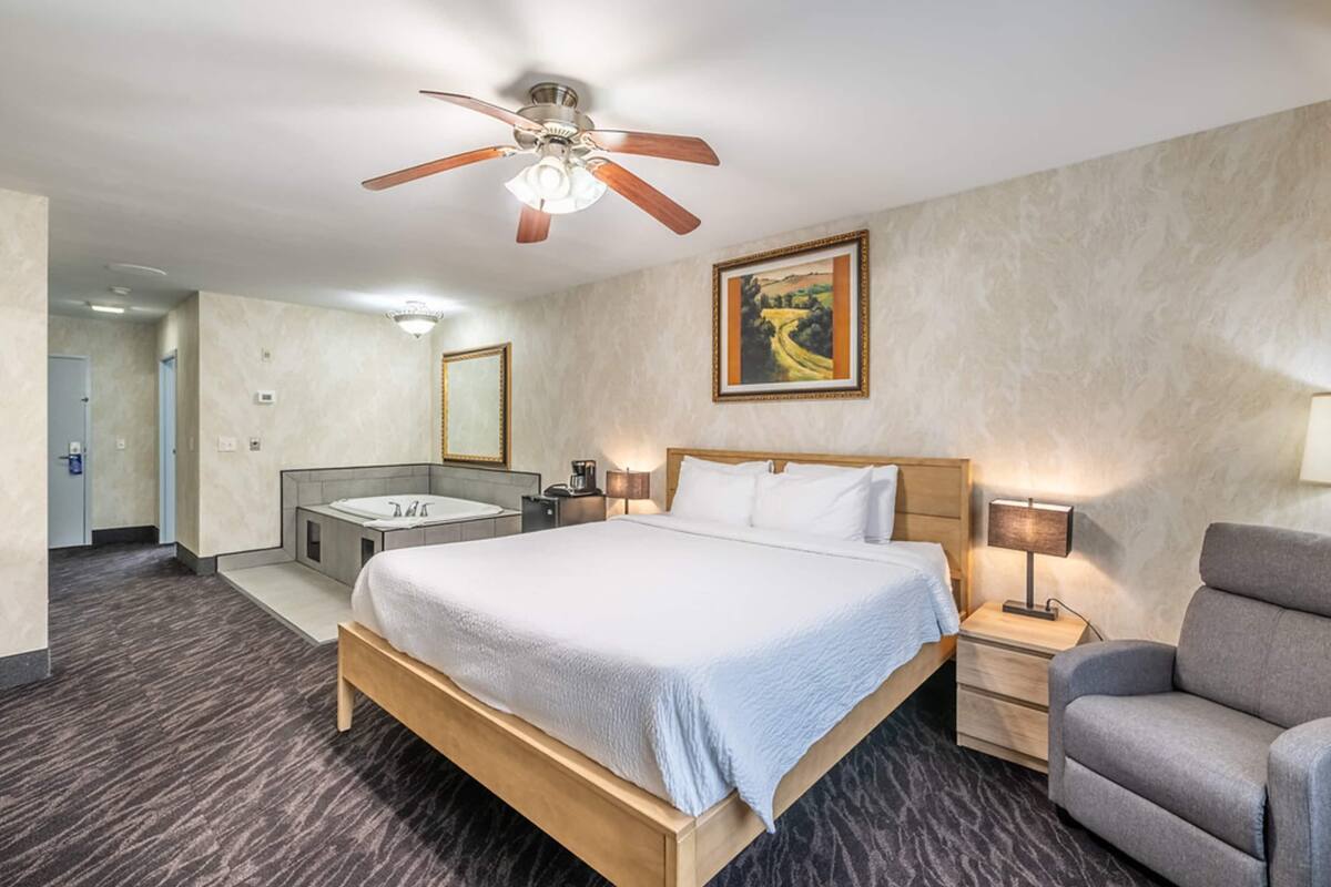 A hotel room with a bed, a desk, a chair, a ceiling fan, a painting, and a bathroom with a bathtub.