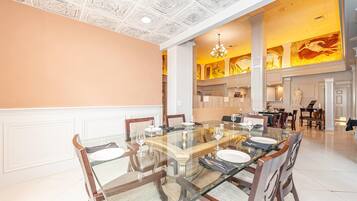 A spacious dining area with a large glass table surrounded by wooden chairs, white walls, and a high ceiling with intricate designs.