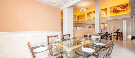 Un vaste espace dédié à la salle à manger, avec une grande table en verre entourée de chaises en bois, des murs blancs et un plafond haut orné de motifs complexes.