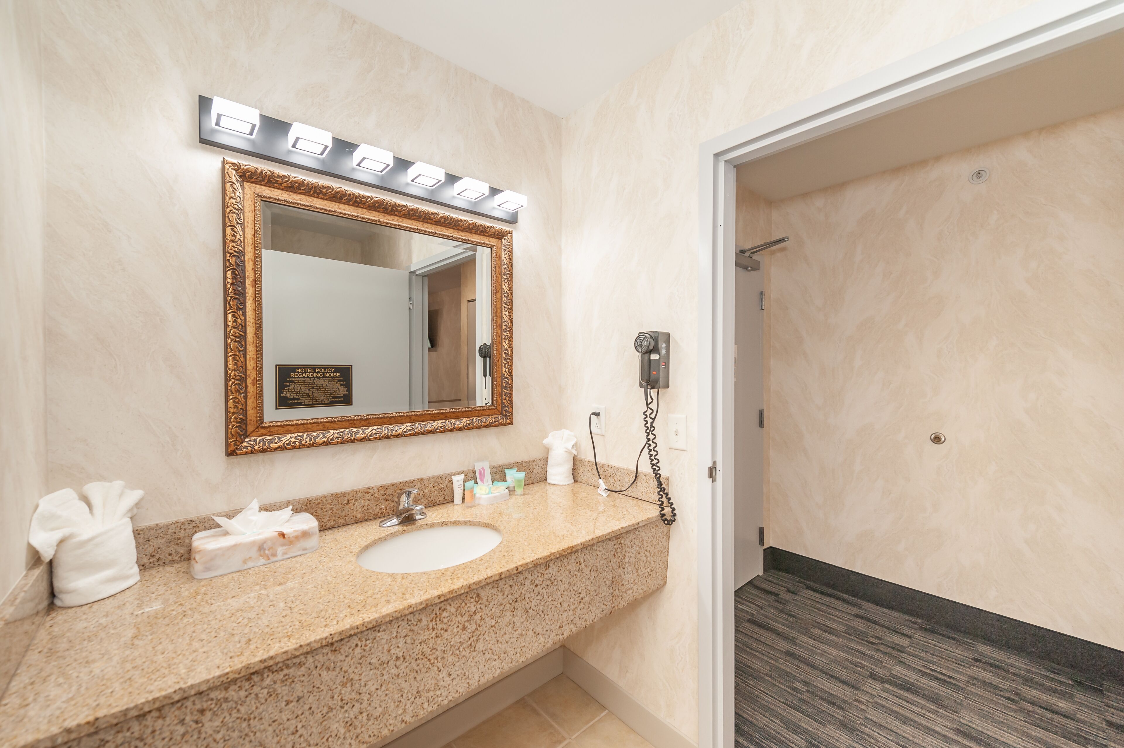 A bathroom with a marble sink, a large mirror with lights, a handheld showerhead, and a towel dispenser.