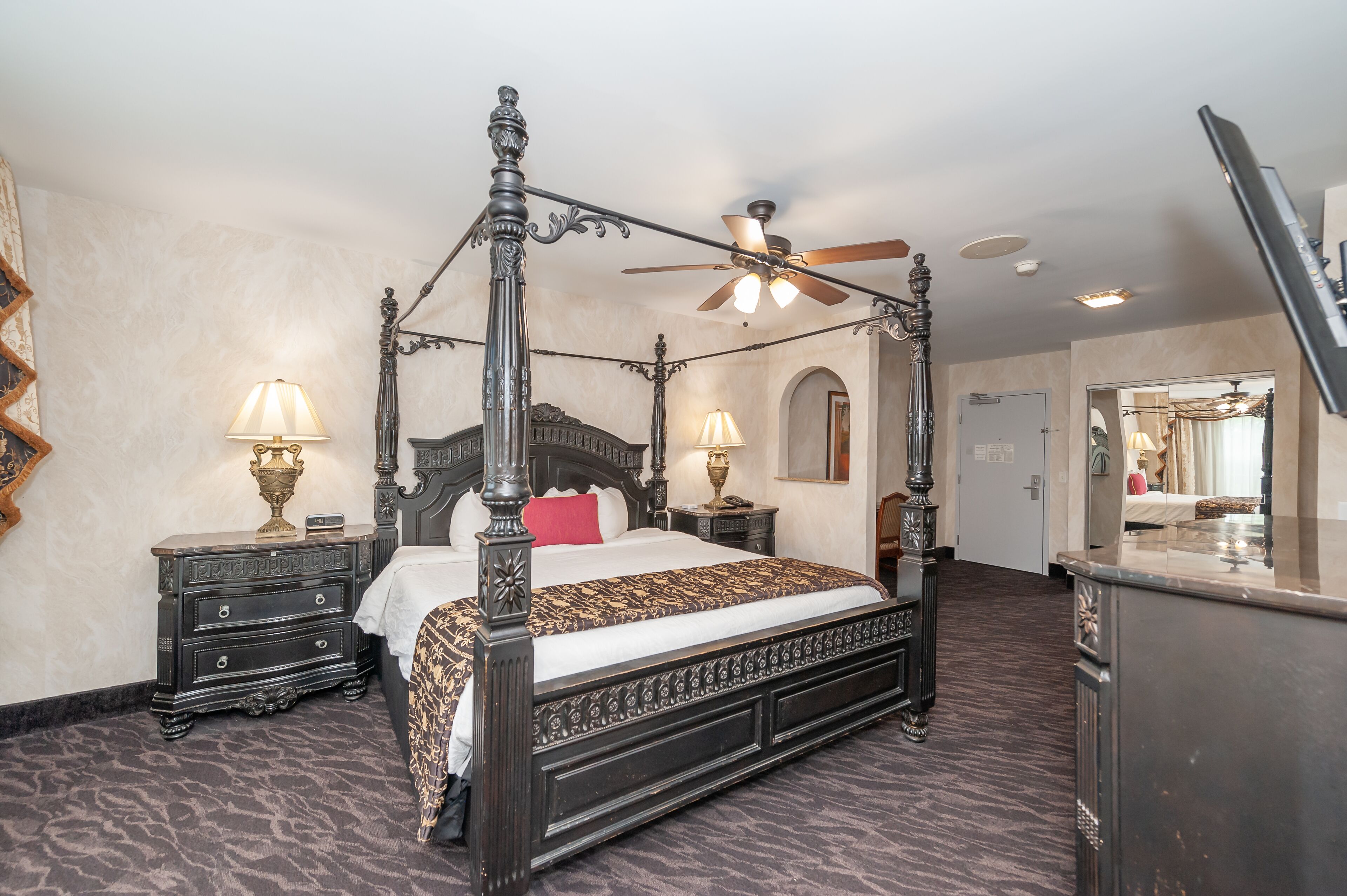 A four-poster bed with ornate metalwork, two bedside tables with lamps, a ceiling fan, and a marble-topped dresser.