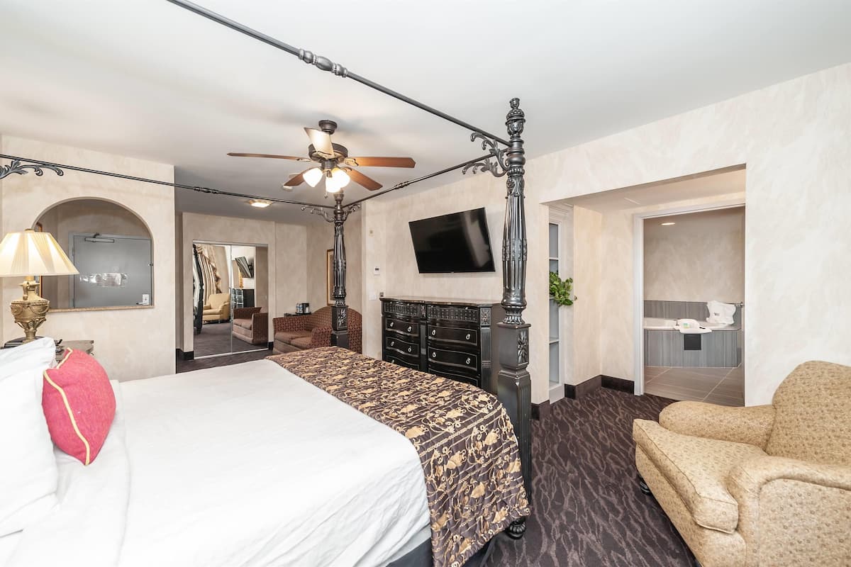 A hotel room with a four-poster bed, a ceiling fan, a flat-screen TV, a desk with a lamp, and a sofa.