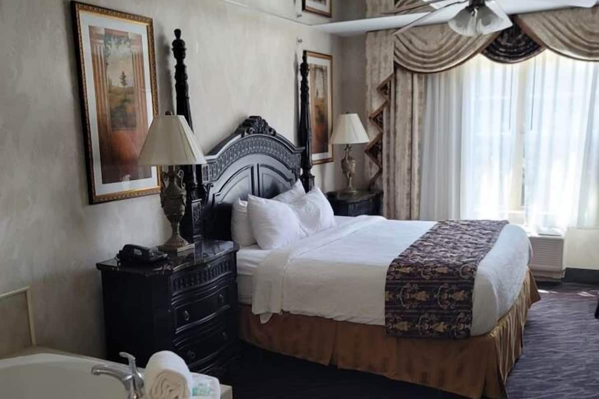 A hotel room with a large bed, a separate bathroom with a bathtub, a ceiling fan, and a window with curtains.