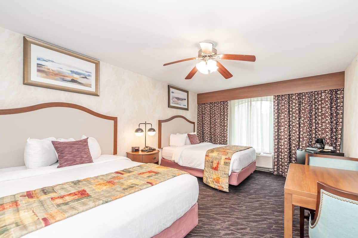 A hotel room with two beds, a ceiling fan, a desk with a chair, and a window with patterned curtains.