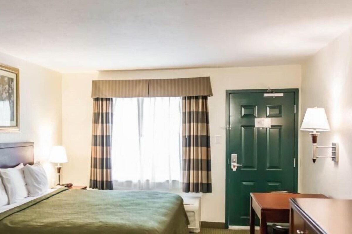 Una habitación de hotel con cama, escritorio, silla, puerta y ventana con cortinas.