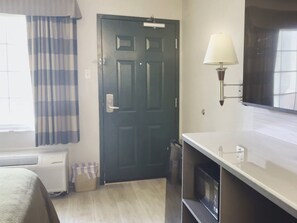 Una habitación de hotel con una puerta oscura, una ventana con cortinas, una lámpara de pared, un televisor de pantalla plana y un radiador.