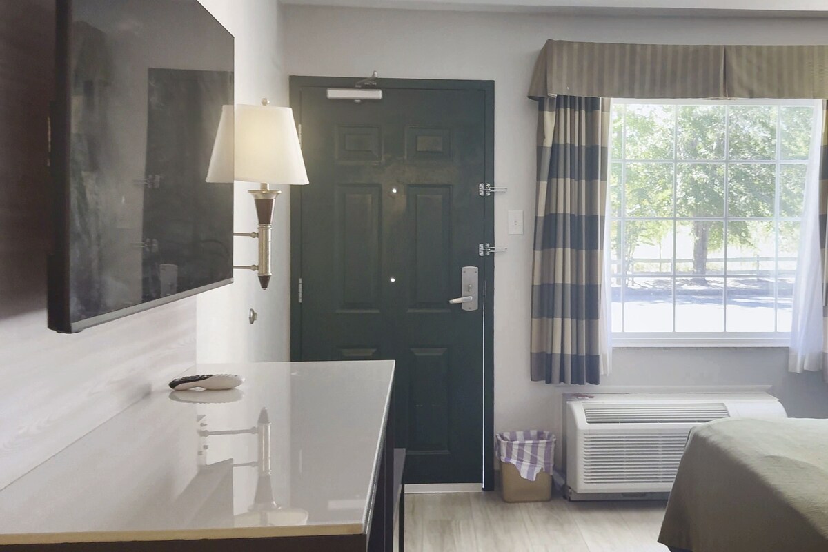 Habitación de hotel con cama, escritorio con lámpara, puerta, ventana con cortinas y unidad de aire acondicionado.