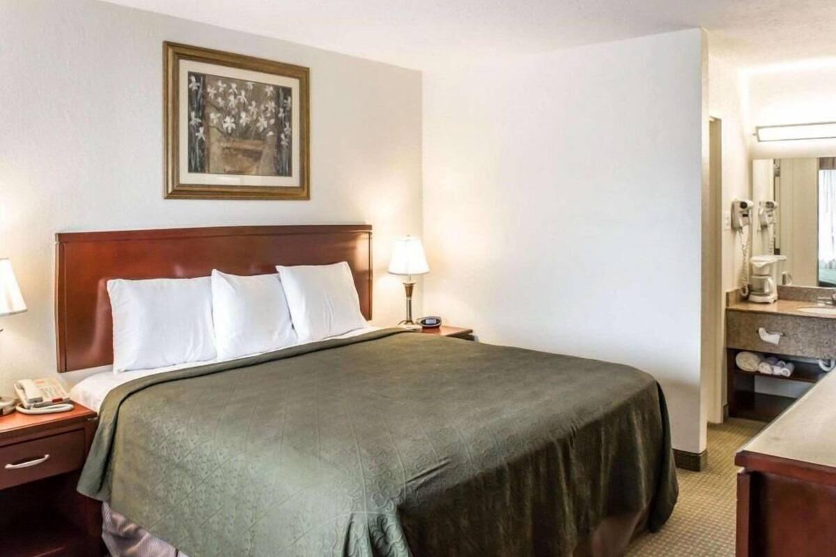 A hotel room with a bed, a nightstand with a phone, a lamp, a painting on the wall, and a bathroom visible through an open door.