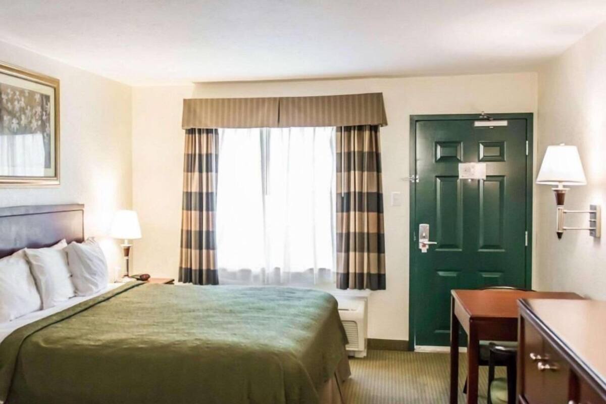 A hotel room with a bed, a desk, a chair, a door, and a window with curtains.