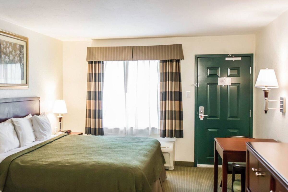 A hotel room with a bed, a desk, a chair, a door, and a window with curtains.