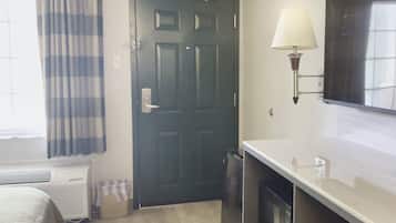A hotel room with a dark door, a window with curtains, a wall-mounted lamp, a flat-screen TV, and a radiator.