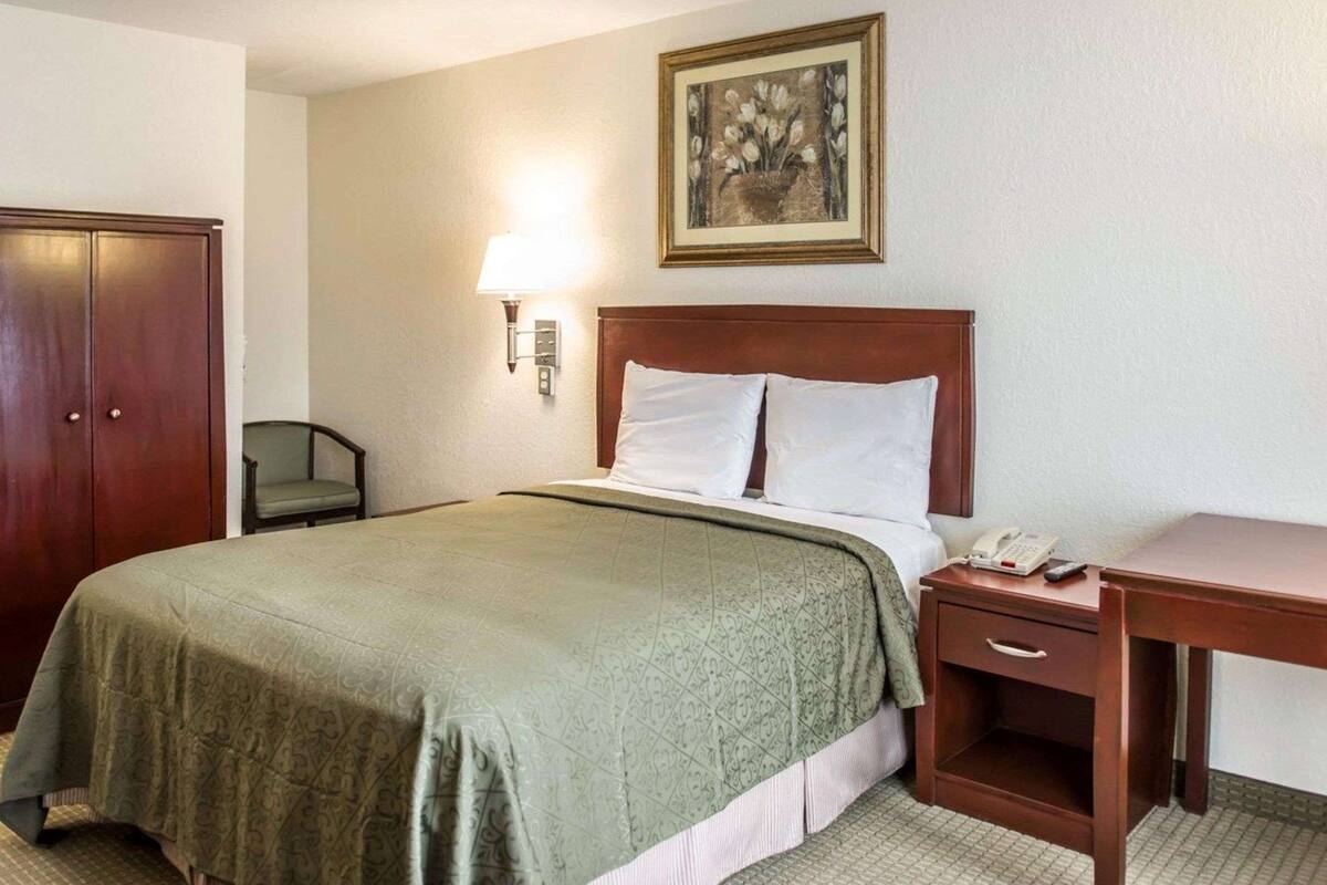 A hotel room with a bed, a nightstand with a phone, a desk, a chair, and a wardrobe.