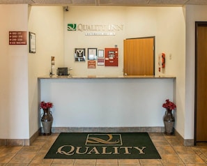 A reception area with a sign for Quality Inn, a reception desk, a fire alarm, and a doormat with the word 'QUALITY'. - Quality Inn Durand I-69 (Durand)