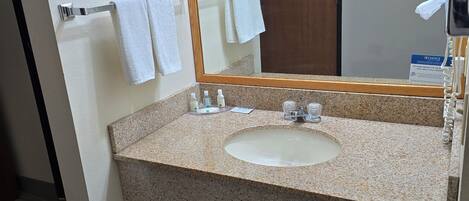 Un baño con lavamanos de encimera de granito, un espejo, toallero con toallas blancas, un cesto de basura y un secador de pelo.