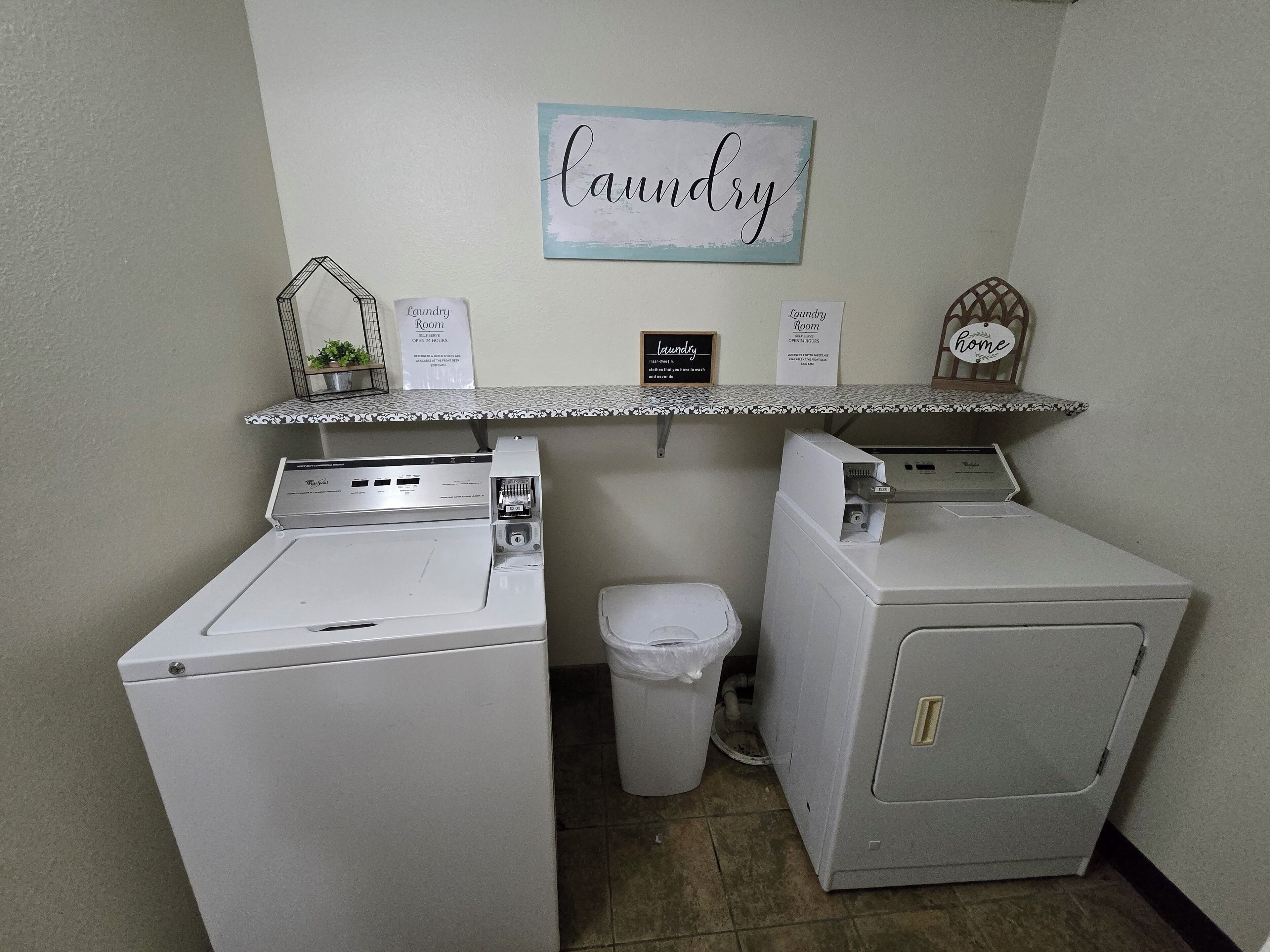 A laundry room with two washing machines, a trash bin, and a shelf with signs indicating the room's purpose and operating hours.