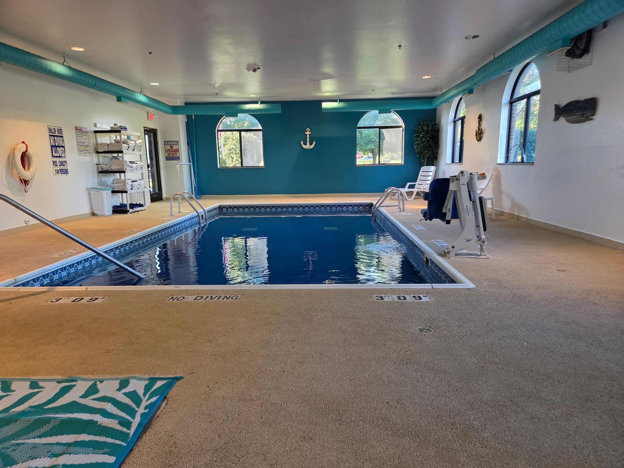 Una piscina interior con un cartel de prohibición de clavados, una silla de salvavidas y un letrero de capacidad de la piscina.