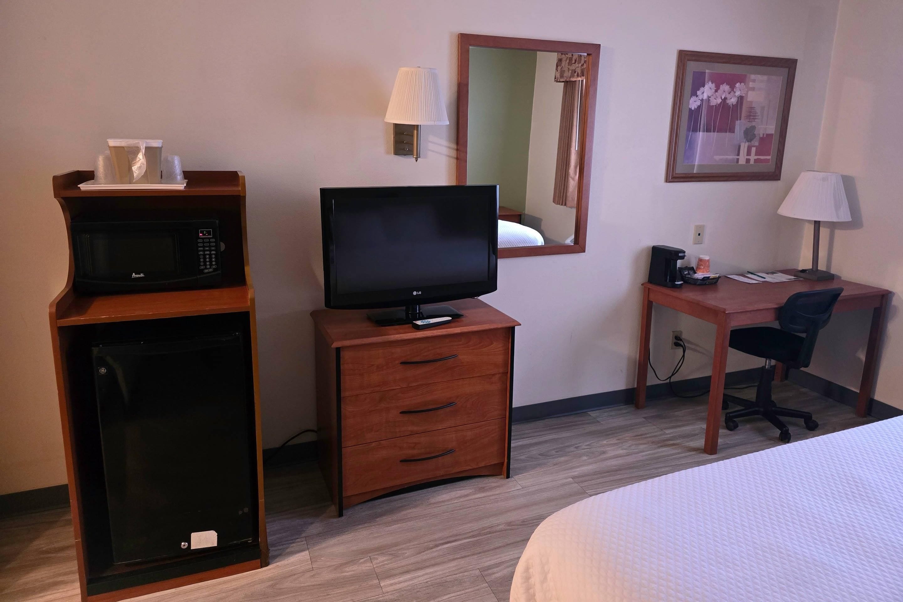 Una habitación de hotel con una cama, un escritorio con una silla, un televisor, un microondas, una pequeña mesita de noche y una lámpara, además de un cuadro en la pared.