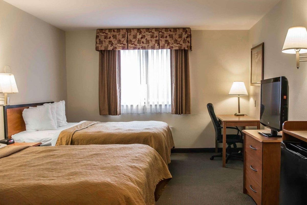 Habitación de hotel con dos camas, un escritorio con computadora, un televisor y una ventana con cortinas.