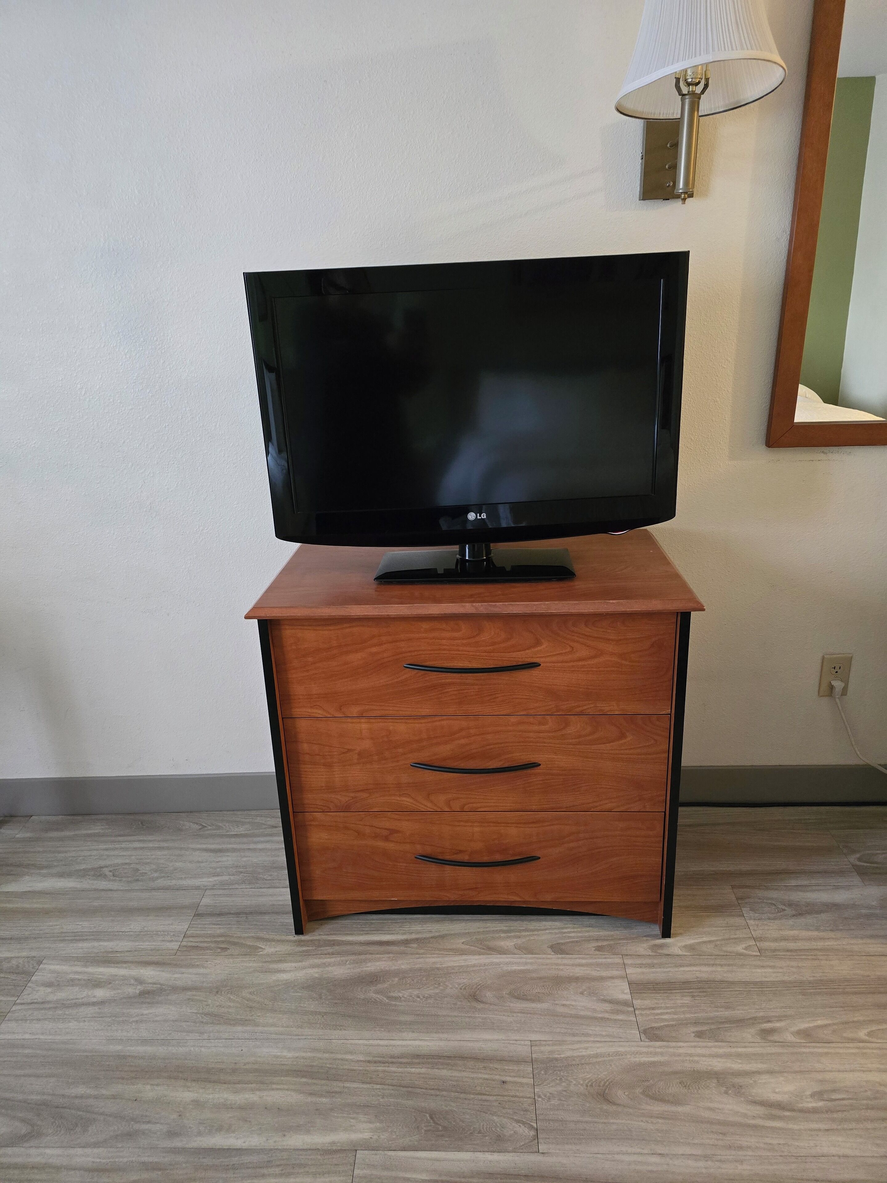 A flat-screen TV on a wooden cabinet with three drawers, a lamp on the wall, and a power outlet.