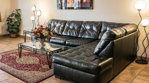 Ein Wohnzimmer mit einem schwarzen Leder-Modulsofa, einem Glastisch mit floralem Mittelstück, einem rot gemusterten Teppich und einem Bild an der Wand.