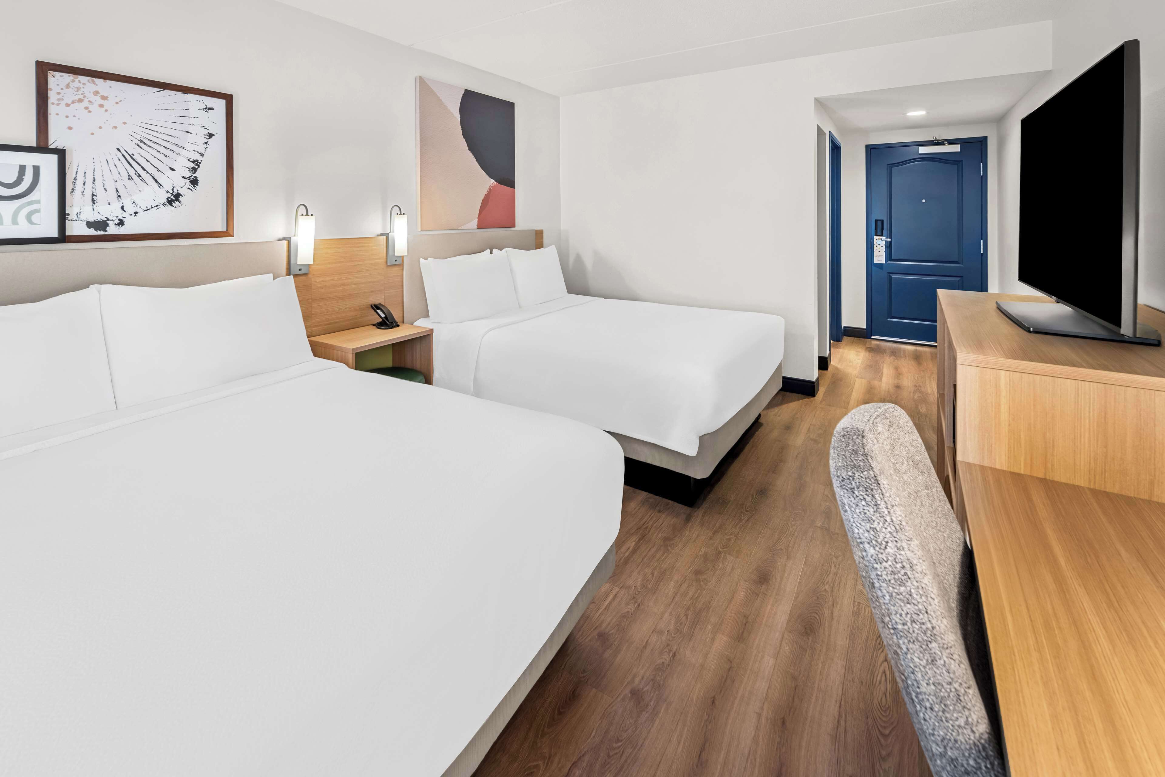 A hotel room with two beds, a desk, a chair, a TV, and artwork on the walls.
