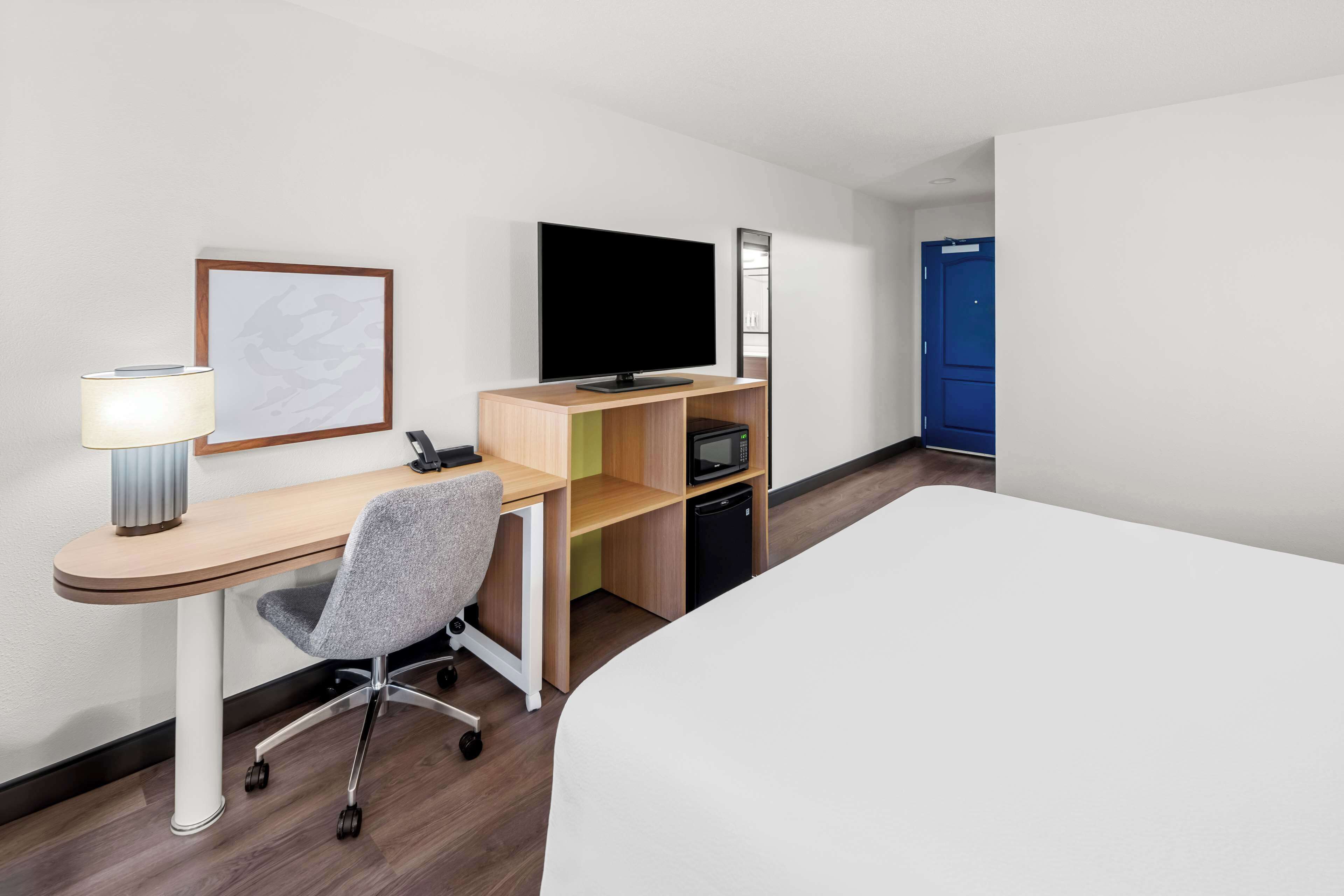 Ein Hotelzimmer mit einem Bett, einem Schreibtisch mit Stuhl, einem Flachbildfernseher, einer Lampe, einer Mikrowelle und einer blauen Tür.