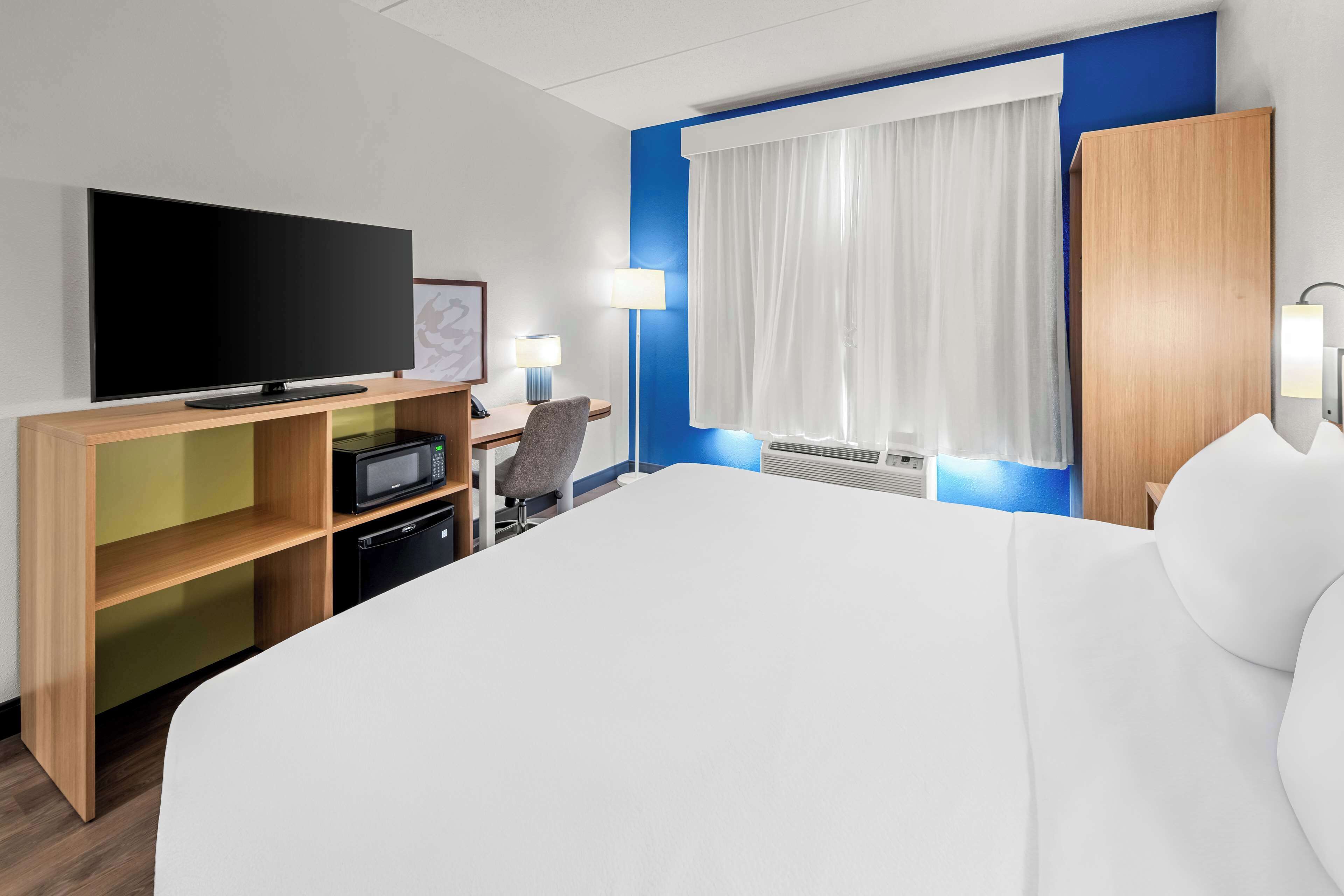 A hotel room with a large bed, a flat-screen TV, a desk with a chair, a microwave, and a window with curtains.