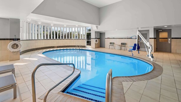An indoor swimming pool with clear blue water, tiled flooring, and a lifeguard chair. - Spark by Hilton Woodbridge (Woodbridge)
