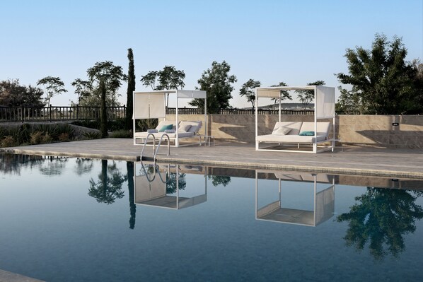 Zona junto a la piscina con dos sillas reclinables blancas bajo pérgolas, rodeada por un muro de piedra y árboles.