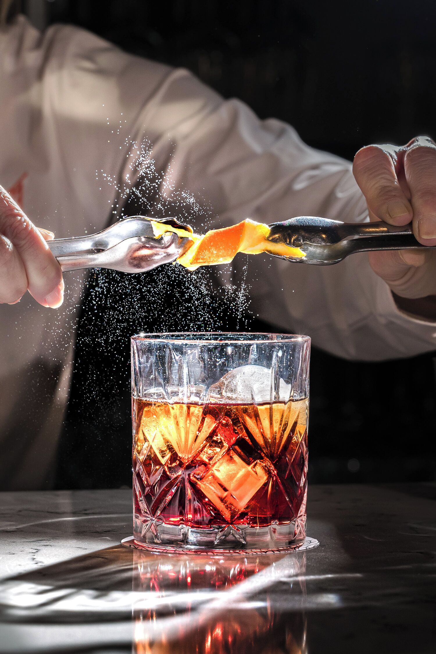 Una persona exprimiendo una naranja en un vaso con líquido ámbar, probablemente un cóctel, con cubitos de hielo.