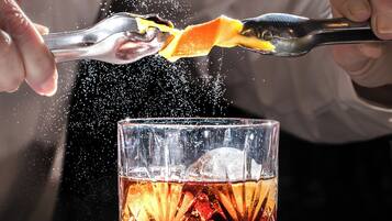 Una persona exprimiendo una naranja en un vaso con líquido ámbar, probablemente un cóctel, con cubitos de hielo.