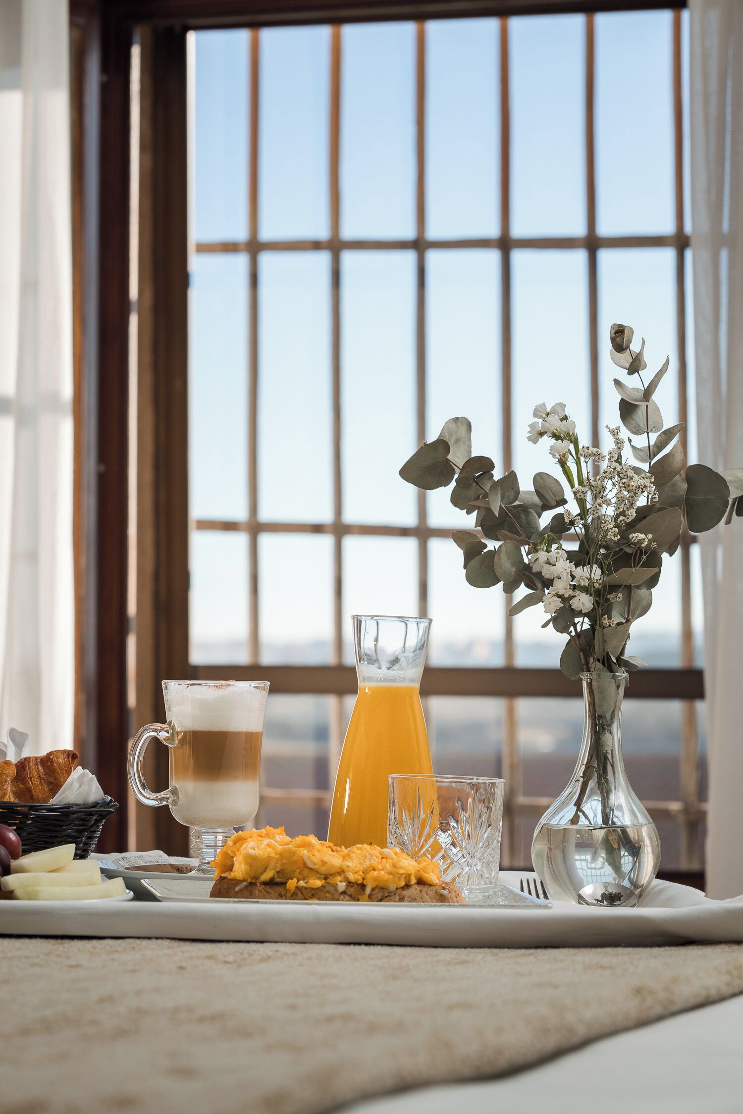 Un desayuno con una taza de café, un vaso de jugo de naranja, huevos revueltos y un florero con flores sobre una mesa.