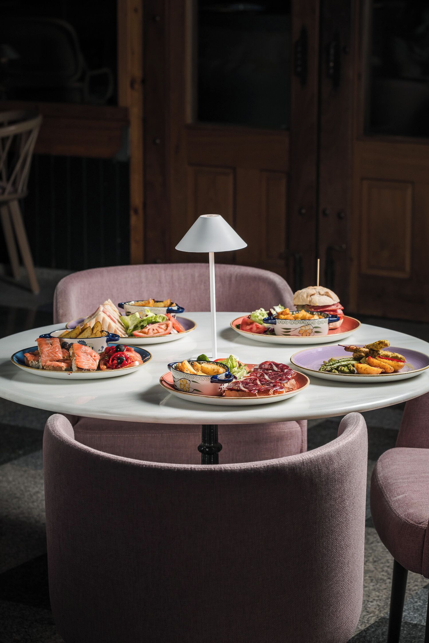Una mesa con varios platos, una lámpara y sillas en una habitación con puertas de madera y una silla al fondo.