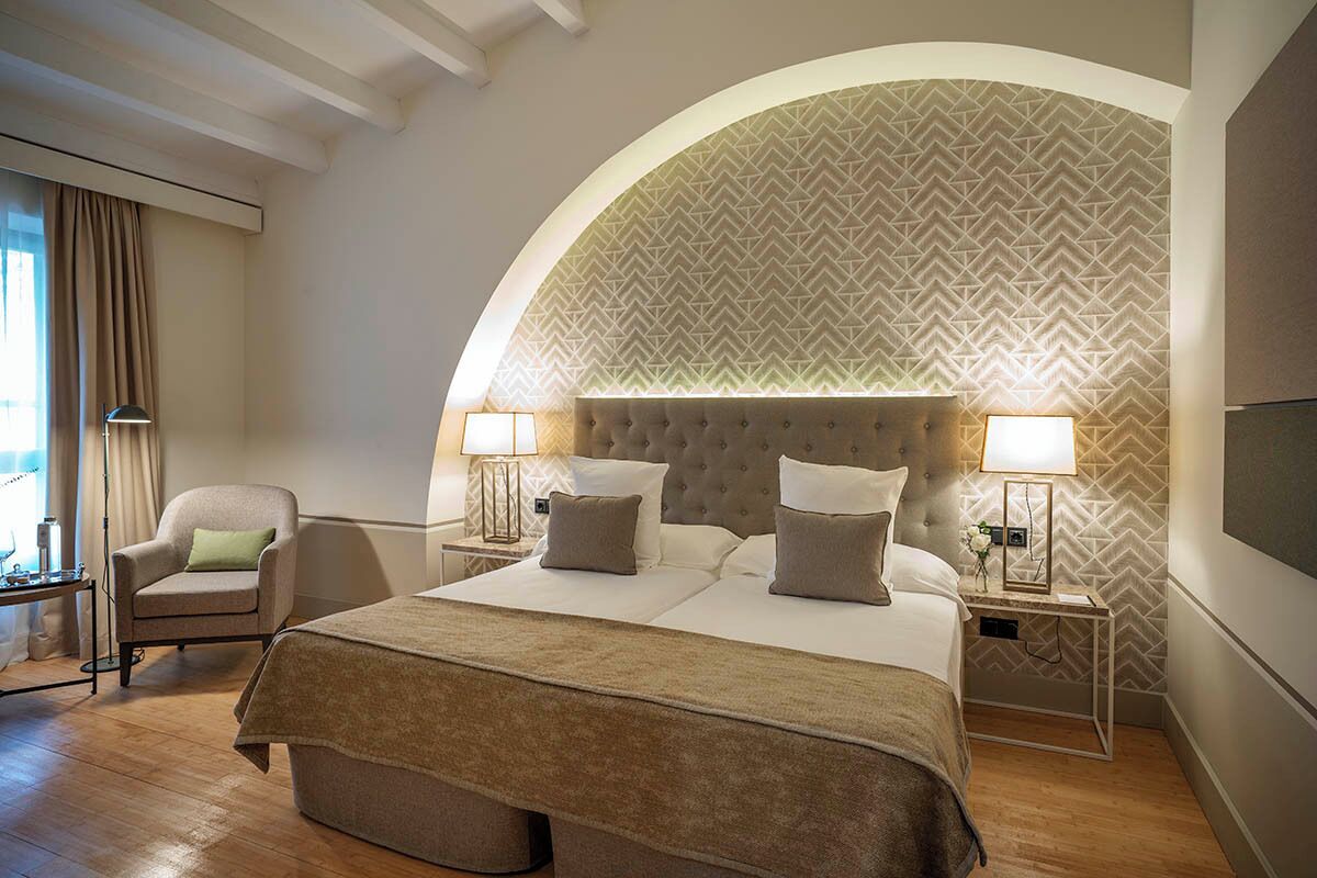 Een hotelkamer met een groot bed, twee nachtkastjes met lampen, een stoel en een gebogen muur met een geometrisch patroon.