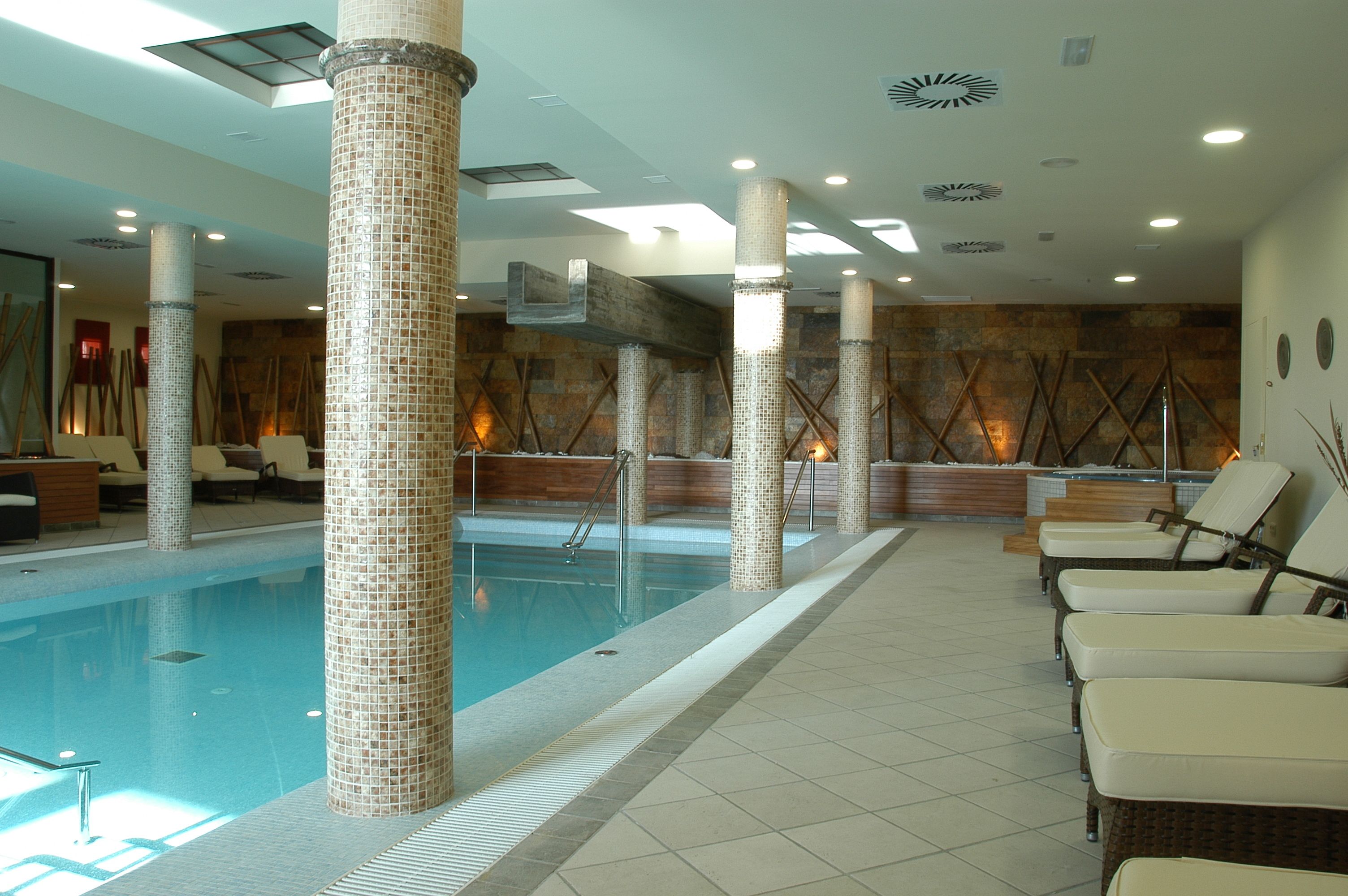 A swimming pool with tiled columns, lounge chairs, and a stone wall with wooden beams.